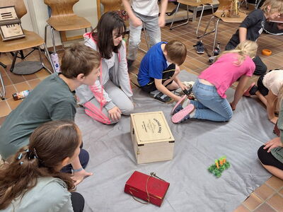 Schüler sitzen am Boden auf einem grauen Tuch, darauf steht eine Holzkiste