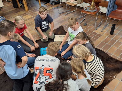 Schüler sitzen versammelt am braunen Fliesenboden auf einem Fell