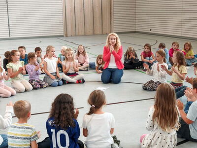 Schulkinder sitzen klatschend auf dem Boden der Turnhalle